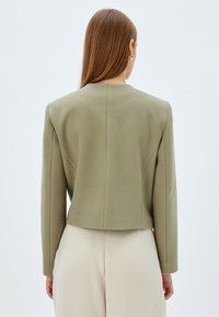 Olivgrüner, taillierter Blazer mit strukturierten Schultern und langen Ärmeln, der eine glatte Textur und klare Linien aufweist. Rückansicht, kombiniert mit cremefarbenen Hosen.