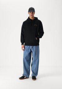 Sudadera negra con capucha, bolsillo frontal y logo naranja, combinada con unos vaqueros de denim azul sueltos y zapatillas negras con suelas marrones.