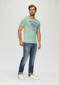 Muntgroene katoenen T-shirt met blauwe bedrukte tekst en graphics, gecombineerd met blauwe denim jeans en witte sneakers. Casual pasvorm, korte mouwen.