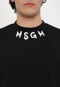Homme portant un sweat à col rond noir avec les lettres blanches et épaisses MSGM imprimées autour du col.