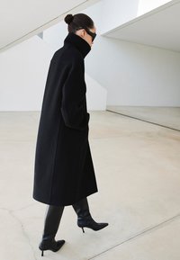 Czarny wełniany płaszcz z wysokim kołnierzem, dużymi kieszeniami i luźnym krojem. Noszony z błyszczącymi czarnymi wysokimi kozakami sięgającymi do uda oraz prostokątnymi okularami przeciwsłonecznymi.