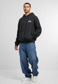 Felpa nera con tasca frontale, caratterizzata da un logo bianco. Indossata con jeans blu larghi e sneaker bianche su uno sfondo neutro.