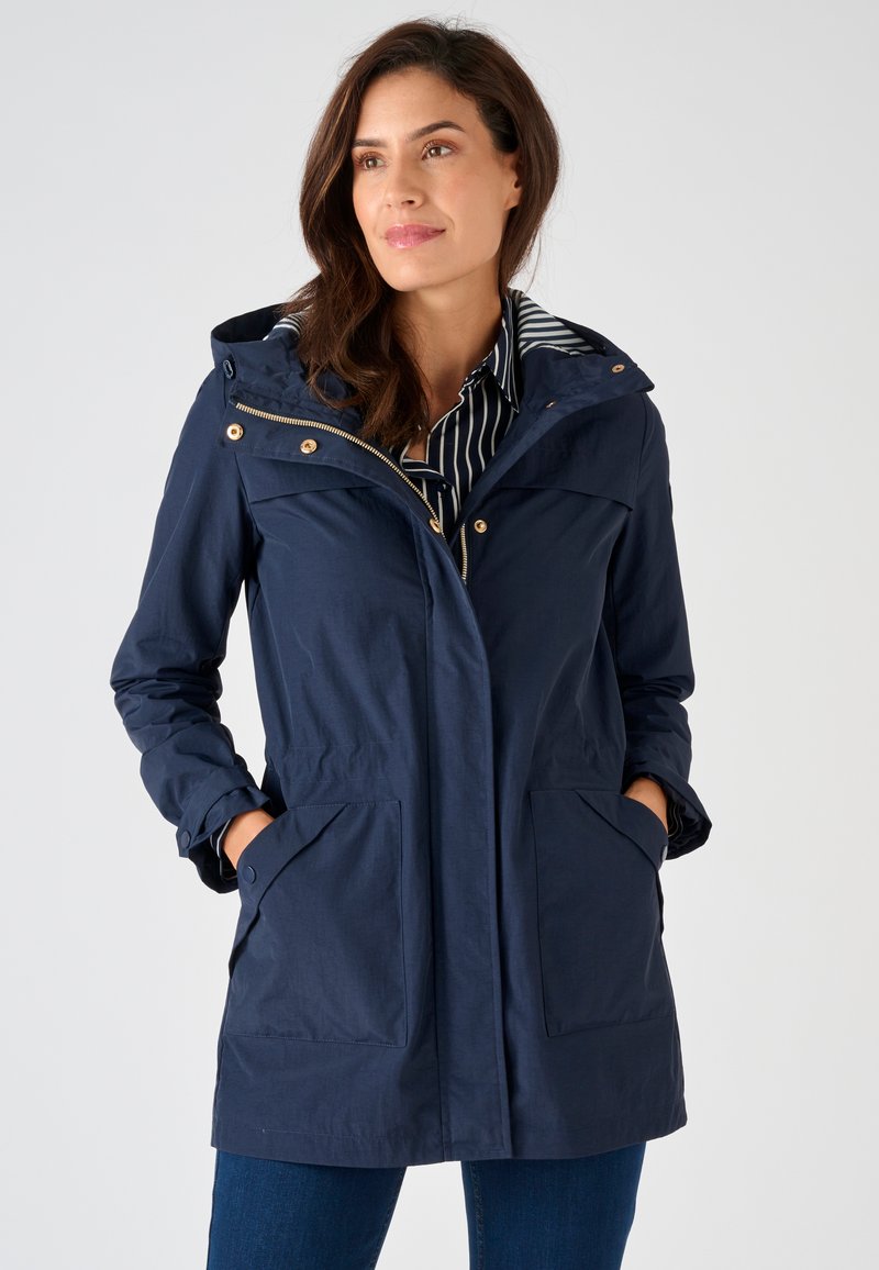 Donna con giacca blu scuro con cappuccio sopra una maglietta a righe, mani in tasca, che guarda leggermente di lato, su sfondo chiaro e uniforme.