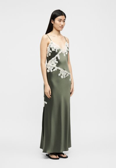 Γυναίκα που φοράει ελαιί πράσινο σατέν φόρεμα με λεπτομέρειες από λευκή φλοράλ δαντέλα και λεπτές τιράντες, στέκεται με τα χέρια χαλαρά στα πλευρά.