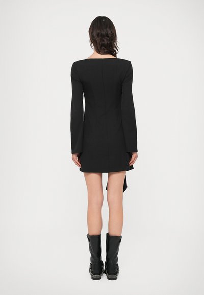 Čierne šaty s dlhým rukávom, štvorcovým výstrihom, prispôsobeným strihom a asymetrickým lemom. Nosia sa s čiernymi čižmami po kolená s prackami.