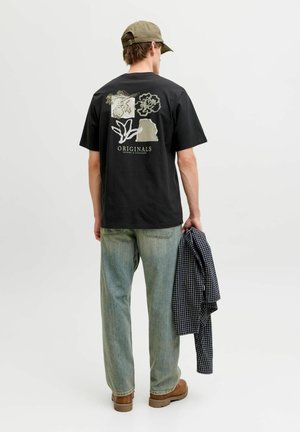 Mężczyzna w czarnej koszulce z nadrukiem kwiatowym i abstrakcyjnym, jasnoniebieskich dżinsach, brązowych butach, oliwkowej czapce, trzymający złożoną koszulę w kratę.