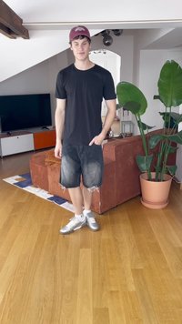 Camisa preta, calções de ganga desgastados e ténis prateados. Quarto com chão em madeira, sofá e plantas de interior num ambiente luminoso.