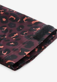 Kokkupandud varrukas kirjaga, mis on tumepunase ja musta leopardimustriga kangas, millel on must Velcro rihma sulgemine valgel taustal.