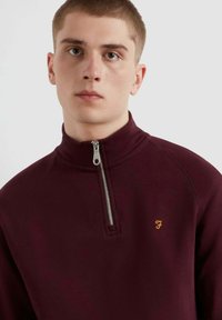 Felpa bordeaux con zip a quarti realizzata in tessuto morbido, caratterizzata da una cerniera argentata e un piccolo logo dorato sul petto.