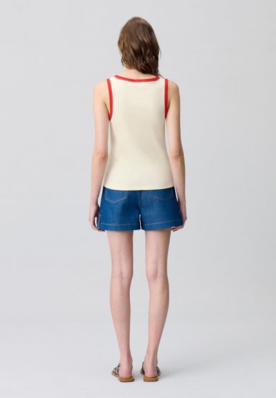 Top a canottiera crema con bordi rossi, abbinato a pantaloni shorts in denim blu. Design semplice, senza maniche, con dettagli sulle cuciture posteriori. Scarpe basse.