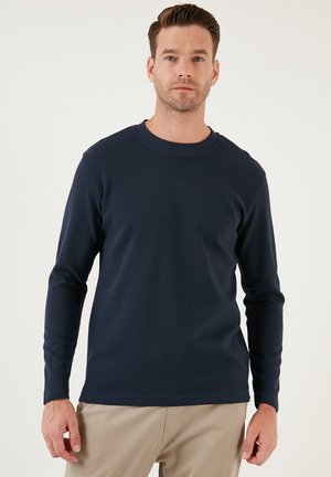 Mornarska košulja s dugim rukavima od teksturirane tkanine, s okruglim izrezom i uskim rukavima, uparenim s bež hlačama. Minimalistički dizajn, bez uzoraka.