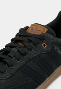 Κοντινό πλάνο μαύρου αθλητικού παπουτσιού Adidas Samba με μαύρα κορδόνια, καφέ δερμάτινο λογότυπο και σόλα από καουτσούκ χρώματος καραμέλας.