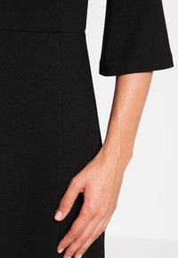 Czarna sukienka z teksturowanej tkaniny, z rękawami dzwonowymi ¾. Zbliżenie na rękę opartą na sukience, podkreślające szczegóły szwu.