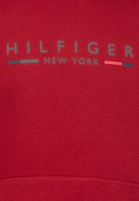 Raudonas medvilnės džemperis su užrašu "HILFIGER NEW YORK" pilkame fone ir spalvotais akcentais. Lygi tekstūra ir banguotas hem'as.