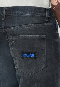 Mørkegrå denimjeans med en baklomme utstyrt med en svart patch som viser den blå logoen "Hugo." Teksturen virker glatt med en subtil fading.