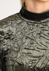 Olivgrüner, strukturierter Stoffoberteil mit hohem Kragen und zerknitterter Oberfläche. Das Material hat ein glänzendes, weiches Erscheinungsbild.