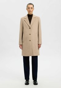 Beige wollen jas met een gesneden kraag, twee voorkeurszakken, drie knopen en een gladde textuur, gecombineerd met donkere broeken en zwarte schoenen.
