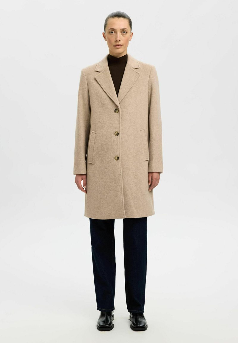 Beige wollen jas met een gesneden kraag, twee voorkeurszakken, drie knopen en een gladde textuur, gecombineerd met donkere broeken en zwarte schoenen.