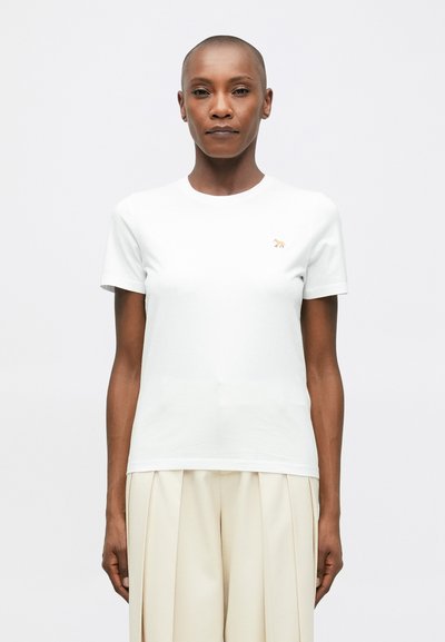 Personne aux cheveux courts portant un t-shirt blanc uni avec un petit logo d'animal et un pantalon beige clair ample à plis, de face.
