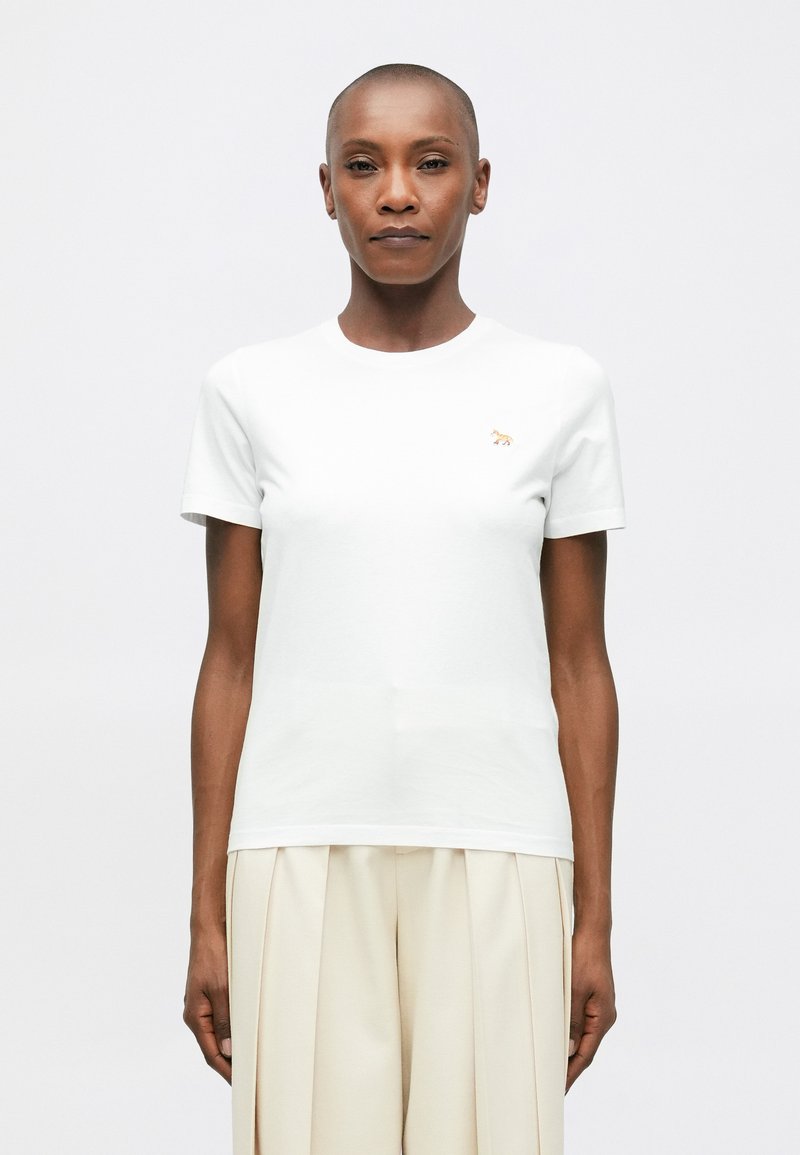 Osoba s krátkými vlasy, oblečená v jednoduchém bílém tričku s malým logem zvířete a volnými světle béžovými plisovanými kalhotami, čelící vpřed.