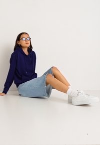 Námornícka modrá mikina s kapucňou a šnúrkou, svetlomodrá džínsová sukňa s surovým okrajom, biele tenisky a zodpovedajúce biele členkové ponožky. Ploché pozadie.