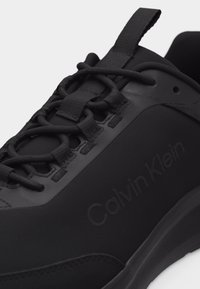 Μαύρο αθλητικό παπούτσι Calvin Klein με στρογγυλά μαύρα κορδόνια, υφασμάτινη θηλιά και χοντρή σόλα, με λεπτομέρεια λογότυπου στο πλάι.