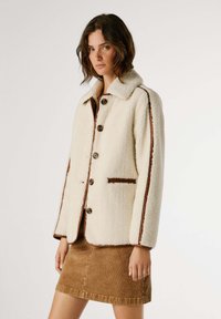 Giacca in shearling color crema con accenti marroni, dotata di collo, bottoni frontali e due tasche laterali.