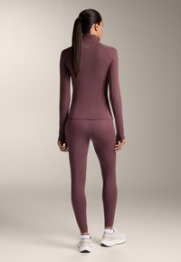 Tajtat aktivklädesset i bordeaux, tillverkat av stretchmaterial. Långa ärmar och hög krage på toppen, parat med smala leggings. Vita sneakers.