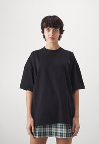Camiseta negra de gran tamaño, tela de algodón, mangas cortas dobladas, escote redondeado, combinada con una falda de cuadros verdes, detalles de dobladillo visibles.