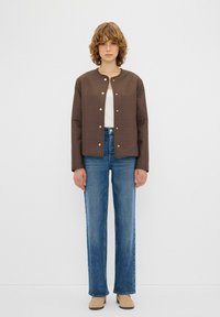 Giubbotto marrone trapuntato con scollo rotondo, bottoni a pressione, indossato sopra una maglietta bianca. Abbinato a jeans blu a vita alta e scarpe beige.