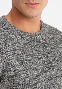 Šedý pletený svetr s kulatým výstřihem, zdobený texturovaným vzorem a žebrovanými manžetami. Materiál působí měkce a pohodlně.