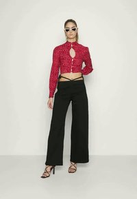 Camisa curta com padrão em vermelho e detalhe em forma de buraco, combinada com calças largas pretas. A modelo usa óculos de sol brancos em forma de gato e sapatos de salto pretos com tiras.