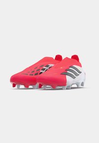 Κόκκινα και άσπρα ποδοσφαιρικά παπούτσια με αεροδυναμικό σχέδιο, που διαθέτουν υφή, μαύρες λεπτομέρειες και μορφοποιημένα πετάλια για πρόσφυση.