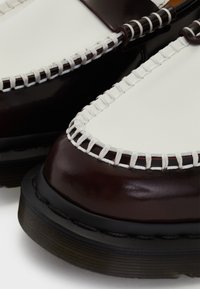 Detailní záběr na hnědou a bílou koženou botu se výrazným bílým prošíváním a silnou černou gumovou podrážkou.