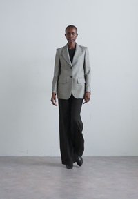 Harmaat kuvioitu bleiseri, jossa on yksi nappi, yhdistetty mustiin leveälahkeisiin housuihin. Sileä pinta ja jämäkkä muotoilu, minimaalinen yksityiskohtaisuus.