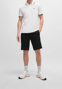 Polo blanc avec des accents noirs, associé à un short noir. Le modèle porte des baskets claires et des chaussettes grises, se tenant devant un fond uni.