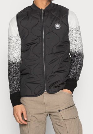 Man wearing a black quilted zip-up vest over a black-and-white speckled long-sleeve sweater paired with beige cargo pants.