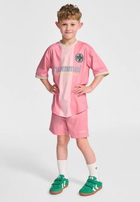 Rosa Sporttrikot mit kurzen Ärmeln und passenden Shorts. Mit einem Logo und hellgrünen Akzenten. Getragen mit grünen Turnschuhen und weißen Socken.