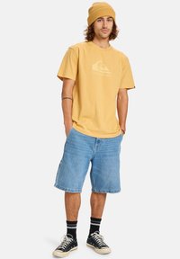 Žluté bavlněné tričko s logem na přední straně; kombinované s světlými modrými denimovými šortkami. Doplňeno béžovou pletenou čepicí a černými teniskami s bílou podrážkou.