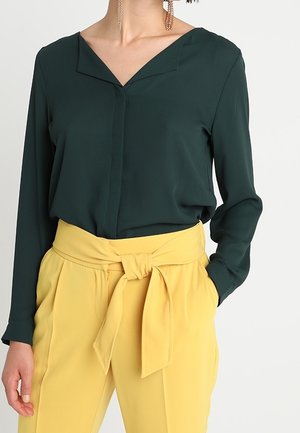 Vrouw draagt een donkergroene blouse met lange mouwen, ingestopt in een hooggesneden gele broek met een strikceintuur van stof.