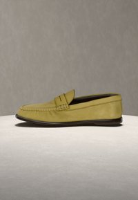 Mocassin en cuir jaune avec semelle noire, surpiqûres apparentes et une petite bride sur le dessus, présenté sur une surface claire.
