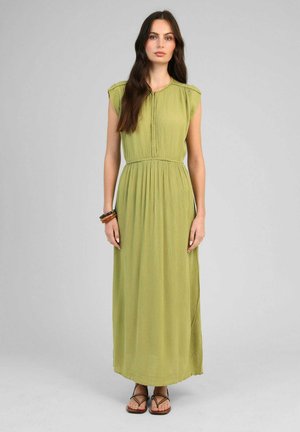 Femme aux longs cheveux bruns portant une robe longue sans manches vert clair, des bracelets marron et des sandales marron à brides, debout devant un fond uni.