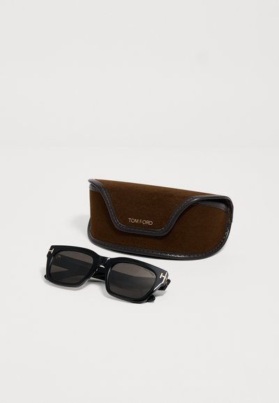 Lunettes de soleil rectangulaires noires Tom Ford avec verres foncés et logo T doré sur les branches, à côté d'un étui Tom Ford en daim et cuir marron.