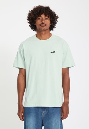 Homme aux cheveux bouclés portant un T-shirt Volcom vert menthe clair et un jean bleu, debout devant un fond blanc.