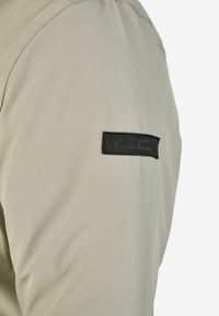 Lichtbeige shirt met korte mouwen en een gladde textuur, met een zwarte rechthoekige stoffen badge op de bovenarm met de tekst "Pierre Cardin."