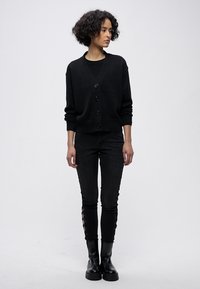 Cardigan negro con escote en V y cinco botones, combinado con jeans ajustados negros con detalles de encaje en los costados. Botines negros.