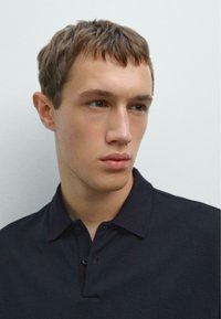Junger Mann mit kurzen braunen Haaren, der ein dunkles Poloshirt trägt und leicht nach links schaut, vor einem schlichten hellen Hintergrund.