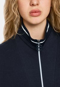 Black zip-up jacket with a high collar, featuring white accents. The zipper has a circular pull tab. Made from a soft, textured fabric.