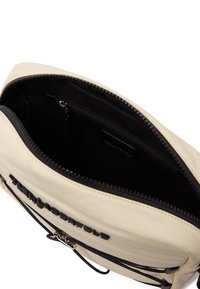 Beige Tasche mit glatter Oberfläche, ausgestattet mit einem schwarzen Reißverschluss und einer inneren Reißverschlusstasche. Das Markenlogo ist auf der Vorderseite geprägt.