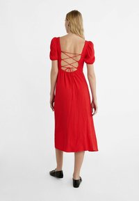 Punainen mekko, jossa on teksturoitu kangas, puffihihat ja ristikkäinen selkäyksityiskohta. Midimittainen, hyvin istuva vyötärö ja laskeutuva hame. Mustat kengät.